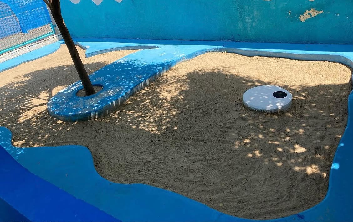 Caja de arena exterior con sombra de árbol y borde pintado de azul