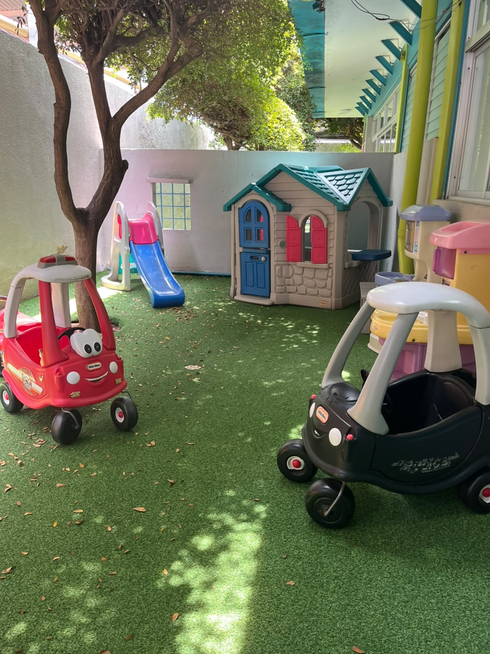 Jardín con grama artificial, casita de juegos, tobogán y carritos a la sombra de un árbol