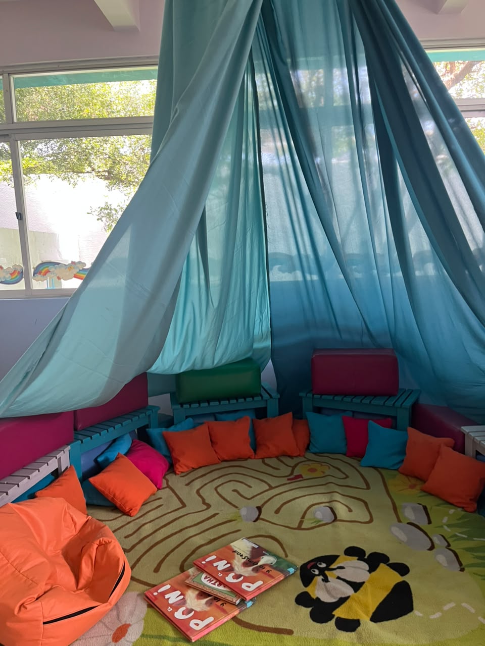 Biblioteca: rincón bajo un dosel de tela con cojines de colores, banquitos y cuentos infantiles sobre la alfombra